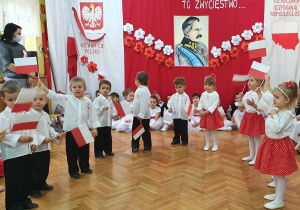 Dziewczynki w stojach biało-czerwonych oraz chłopcy ubrani na galowo stoją w pólkolu, trzymają biało - czerwone chorągiewki i recytują wiersz.